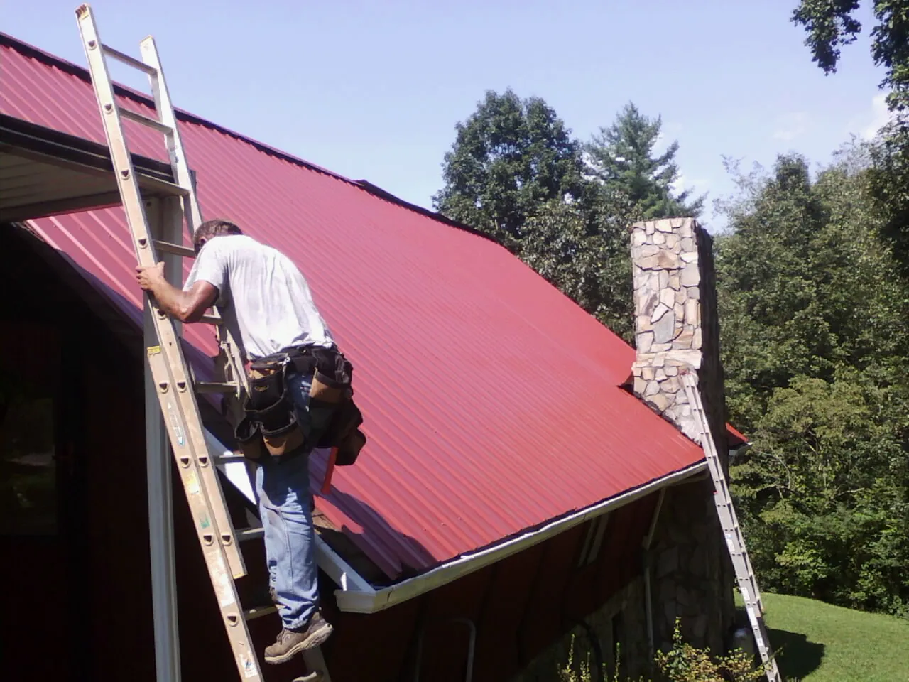 Roofer on a ladder beside a red metal roof and stone chimney — Soffit & Fascia Repair by Roofing Brothers in Glengary